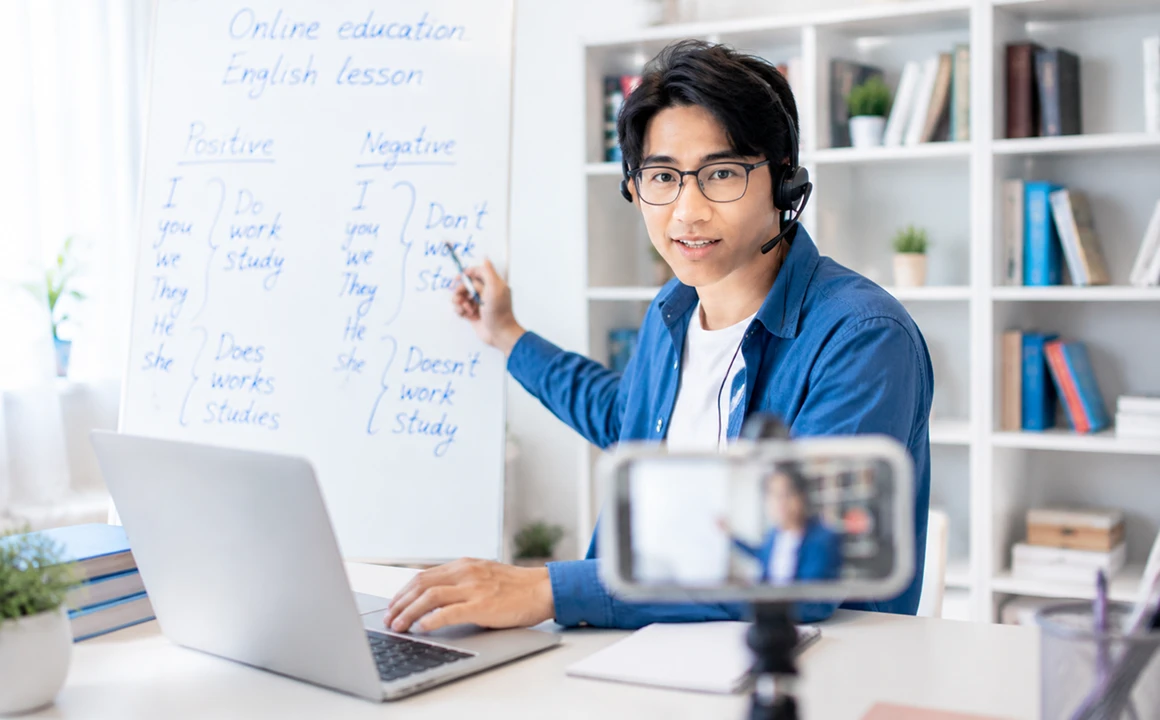 Thầy giáo đeo tai nghe đứng trước bảng trắng viết bài học tiếng Anh, chỉ tay lên bảng, trước mặt là laptop và điện thoại đang quay — ảnh thẻ «Lớp học live tương tác»