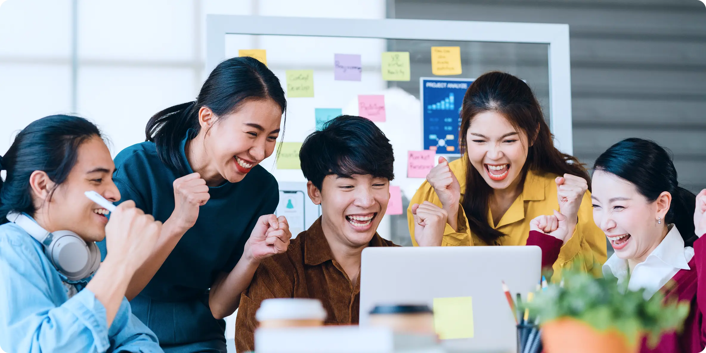 Năm bạn trẻ quây quanh laptop nắm tay reo mừng trước bảng dán giấy ghi chú nhiều màu — ảnh dải «Sứ mệnh của chúng tôi»