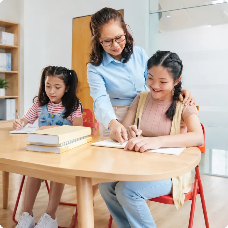 Cô giáo áo sơ mi xanh cúi xuống hướng dẫn hai bé gái viết bài tại bàn gỗ — thẻ «Trường học và Trung tâm đào tạo»