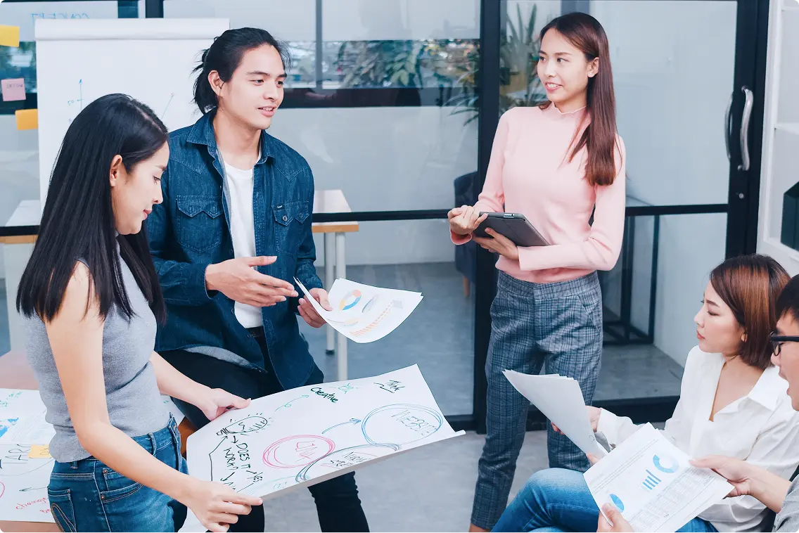 Nhóm năm người trẻ họp trong phòng kính: hai người cầm tờ giấy vẽ sơ đồ ý tưởng, một cô áo hồng cầm máy tính bảng, hai người ngồi xem biểu đồ — ảnh cột phải dải «Lợi ích»