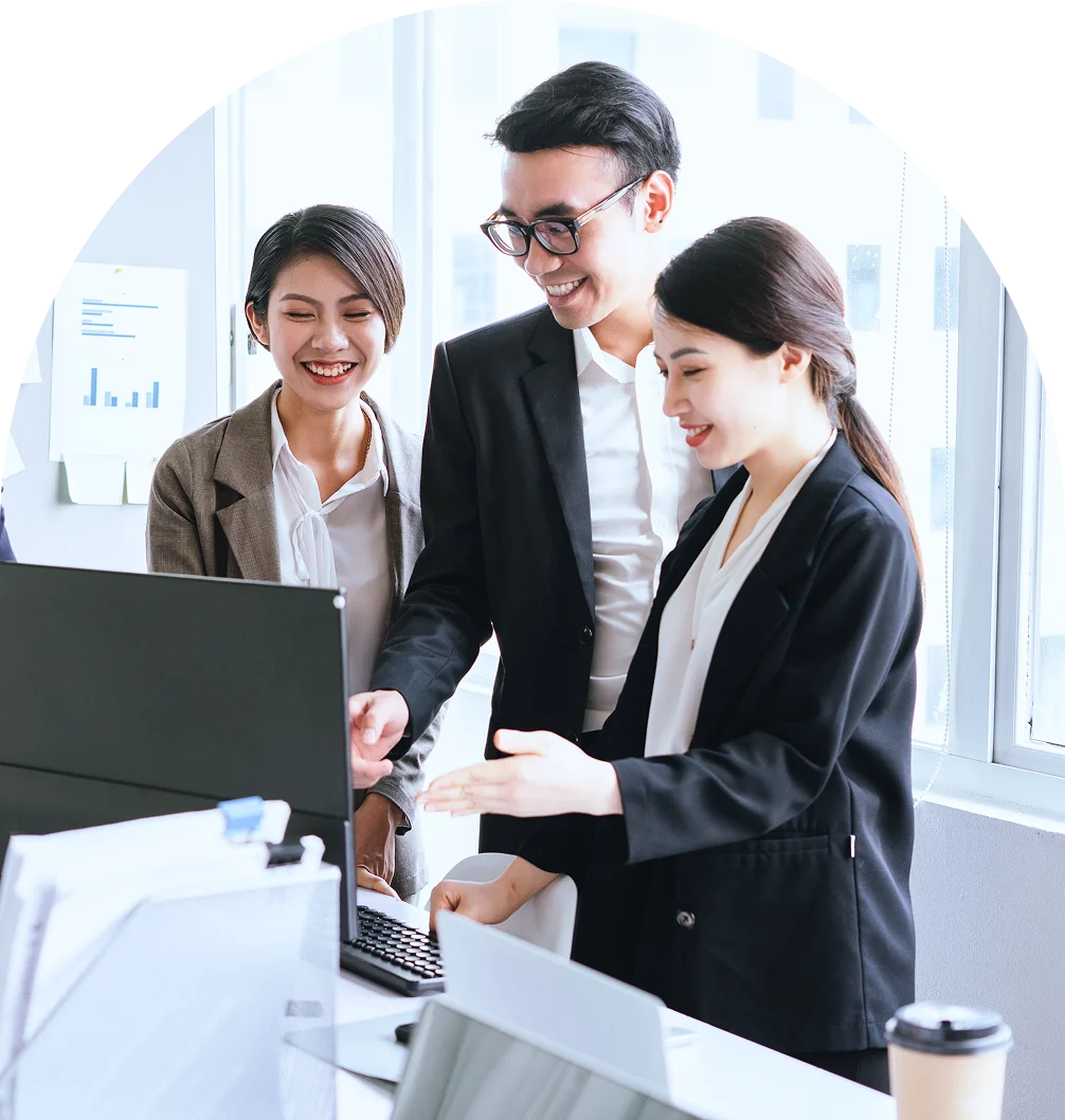 Ba nhân viên văn phòng mặc vest — hai nữ một nam đeo kính — đứng cười bên màn hình máy tính, người nam chỉ tay vào màn hình — ảnh hero trang Đào tạo cho doanh nghiệp